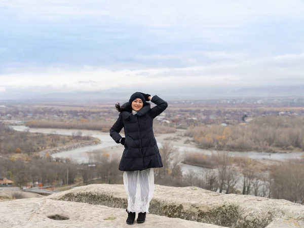 Happy Asian woman tourist in winter cloths standing at the top of Uplistsikhe cave town landmark in Georgia with view of river and sky.