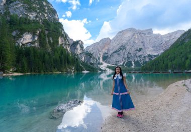 Braies Gölü kıyısında duran mutlu Asyalı kadın turist. Dolomitler, İtalya. Kızla manzara, suda güzel yansıması olan ünlü göl, ağaçlar, bulutlu gökyüzü. Seyahat.