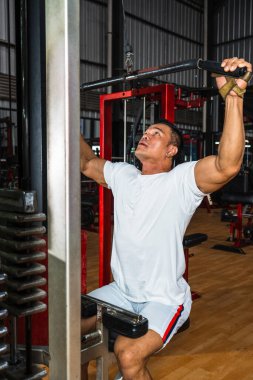 Spor salonunda otururken ağırlık kaldıran beyaz tişörtlü bir sporcunun görüntüsü.