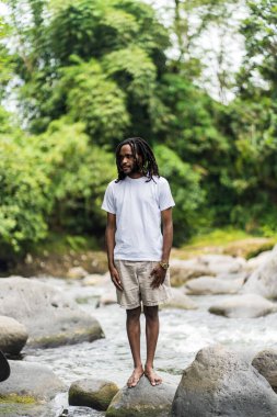 Rastalı, beyaz tişörtlü genç bir adam doğadaki bir nehrin kıyısında dikilirken ayakta dikiliyor.