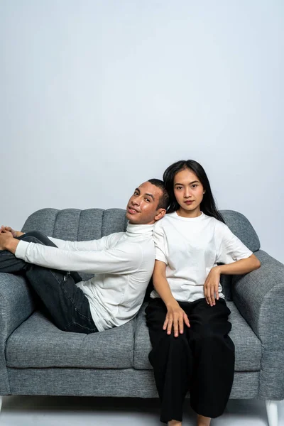 A young couple wearing white blank shirt sitting on a sofa while the man leaning his back to the woman on a white background