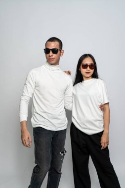 Beyaz gömlekli ve gözlüklü bir çift beyaz arkaplana yan bakarken poz veriyorlar.