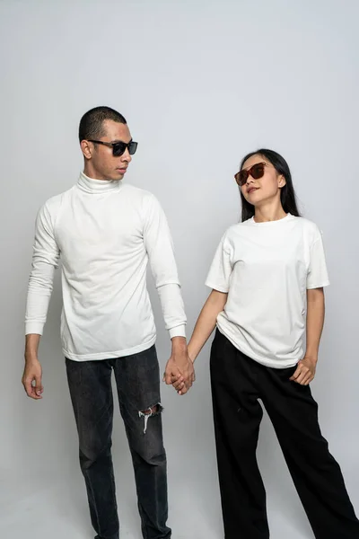 A couple wearing white blank shirt and a glasses looking at each other while holding hands on a white background