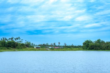Bir sürü ağacın olduğu bir manzara, ıssız bir pavyon, bir göl, ve mavi renkli gökyüzü, görünür bir bulut, renkli bir manzara.