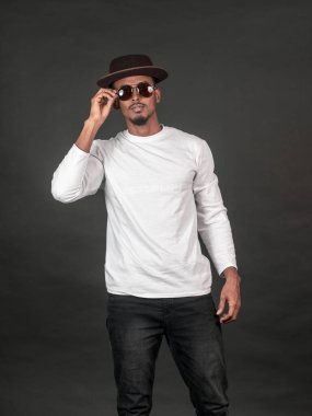 Beyaz gömlekli bir adam siyah arka planda gözlüklerine dokunurken poz veriyor.