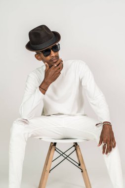 Beyaz gömlekli Afrikalı bir adam, beyaz arka planlı, çenesine dokunurken sandalyenin üstünde oturuyor.