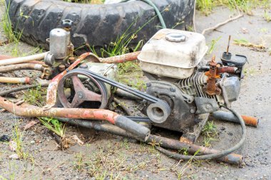 Kırık bir çim biçme makinesi kumlu bir toprağın üzerinde, büyük siyah bir lastiğin yanında duruyordu.