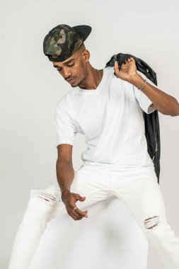 Beyaz v yakalı Afrikalı bir adam beyaz arka planda otururken ve deri ceketini tutarken poz veriyor.