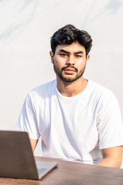 Beyaz tişörtlü, yakışıklı ve sakallı genç bir adam kameraya bakarken dışarıda poz veriyor.