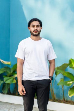 Beyaz tişörtlü genç, yakışıklı, sakallı bir adam kameraya bakarken dışarıda poz veriyor.