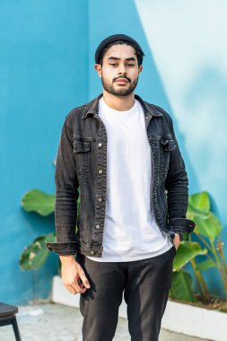 Beyaz tişörtlü genç, yakışıklı, sakallı bir adam kameraya bakarken dışarıda poz veriyor.