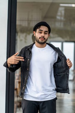 Beyaz tişörtlü genç, yakışıklı, sakallı bir adam kameraya bakarken dışarıda poz veriyor.