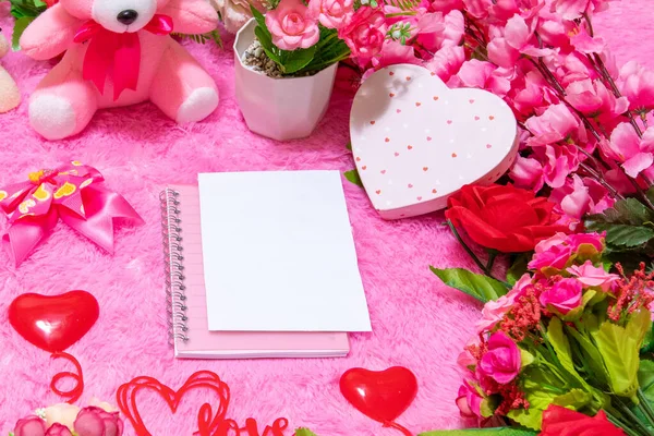 White blank notebook paper on the top of a notebook surrounded by valentine themed decorations, and a pink fluffy carpet as the background