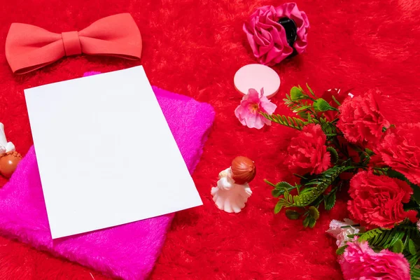 White blank notebook paper above a pink covered notebook surrounded by valentine themed decorations, and a red fluffy carpet as the background