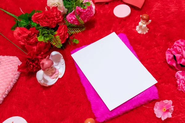 White blank notebook paper above a pink covered notebook surrounded by valentine themed decorations, and a red fluffy carpet as the background