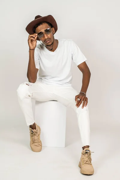 An african man with v-neck shirt sitting on the top of a white box while touching his glasses, with white background