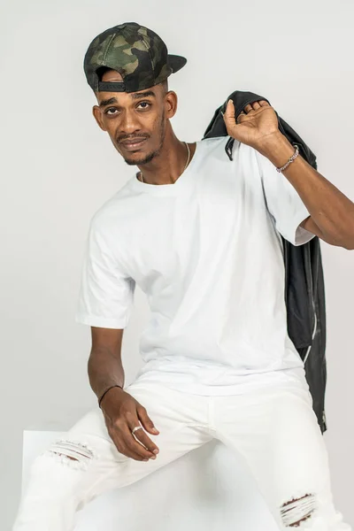 An african man with white v-neck shirt doing a pose while sitting and holding his leather jacket, on the white background