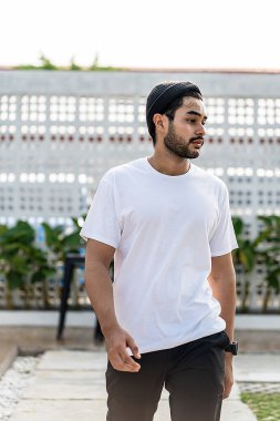 Beyaz tişörtlü genç, yakışıklı, sakallı bir adam dışarıda poz veriyor, yandan bakıyor, tişörtünü taklit ediyor.