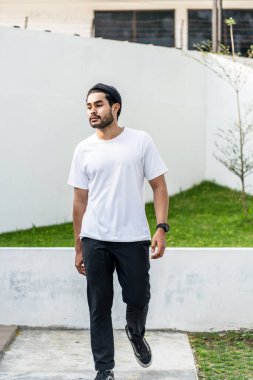 Beyaz tişörtlü genç, yakışıklı, sakallı bir adam dışarıda poz veriyor, yandan bakıyor, tişörtünü taklit ediyor.