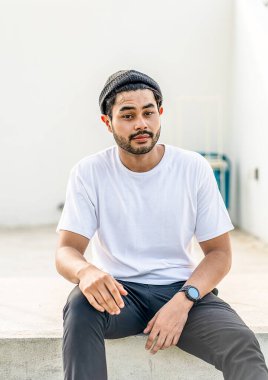 Beyaz tişörtlü, yakışıklı ve sakallı genç bir adam kameraya bakarken dışarıda poz veriyor.