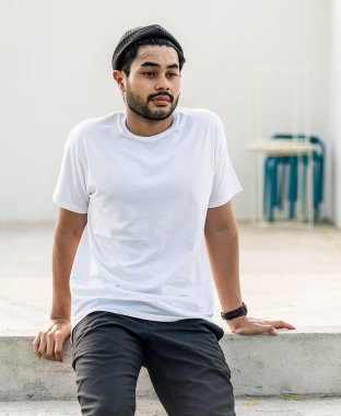 Beyaz tişörtlü genç, yakışıklı, sakallı bir adam dışarıda poz veriyor, yandan bakıyor, tişört süslemesi yapıyor.
