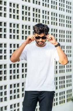 Genç, yakışıklı, sakallı, beyaz tişörtlü bir adam, gözlüklerine dokunurken dışarıda poz veriyor.