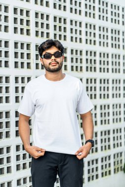 Beyaz tişörtlü genç, yakışıklı, sakallı bir adam kameraya bakarken dışarıda poz veriyor.