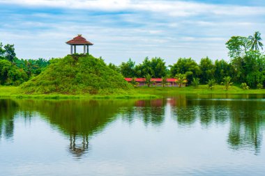 Bir sürü ağacın olduğu bir manzara, ıssız bir pavyon, bir göl, ve mavi renkli gökyüzü, görünür bir bulut, renkli bir manzara.