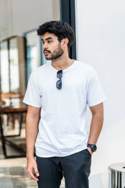 Beyaz tişörtlü genç, yakışıklı, sakallı bir adam dışarıda poz veriyor, yandan bakıyor, tişörtünü taklit ediyor.