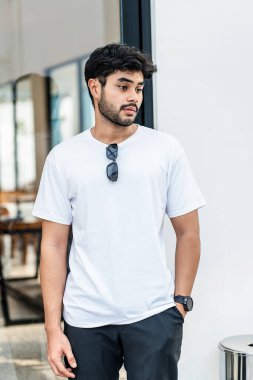 Beyaz tişörtlü genç, yakışıklı, sakallı bir adam dışarıda poz veriyor, yandan bakıyor, tişörtünü taklit ediyor.