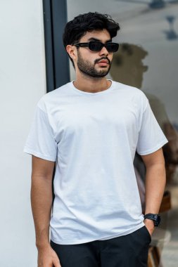 Beyaz tişörtlü genç, yakışıklı, sakallı bir adam dışarıda poz veriyor, yandan bakıyor, tişörtünü taklit ediyor.