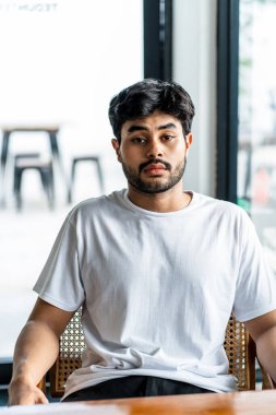 Beyaz tişörtlü genç, yakışıklı, sakallı bir adam kameraya bakarken içeride poz veriyor.