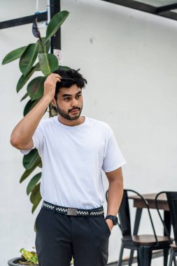 Beyaz tişörtlü genç, yakışıklı, sakallı bir adam dışarıda poz veriyor, yandan bakıyor, tişörtünü taklit ediyor.