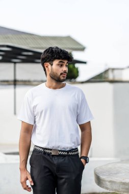 Beyaz tişörtlü genç, yakışıklı, sakallı bir adam dışarıda poz veriyor, yandan bakıyor, tişörtünü taklit ediyor.