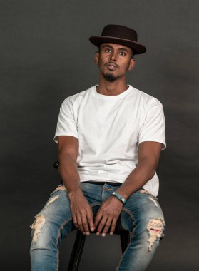 Siyah arka planda, beyaz bir gömlek giyen, poz veren, oturan Afrikalı bir adamın görüntüsü.