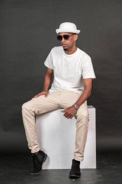 Beyaz bir gömlek giyen Afrikalı bir adam beyaz bir kutunun üstünde oturuyor, siyah arka plan modeli görüntüsü var.