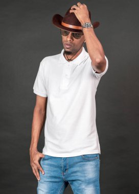 Beyaz tişörtlü, siyah arka planda şapkasına dokunurken poz veren Afrikalı bir adam.