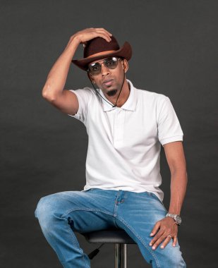 Beyaz, boş polo tişörtlü, gözlüklü ve siyah arka planlı, sandalyenin üstünde oturan bir adam.