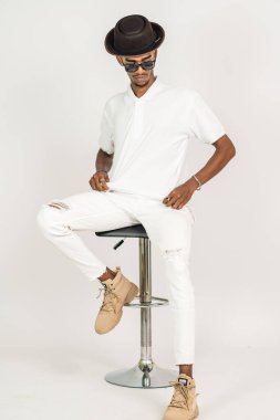 Beyaz, boş tişörtlü, şapkalı ve gözlüklü Afrikalı bir adam beyaz arka planı olan bir sandalyeye oturuyor.