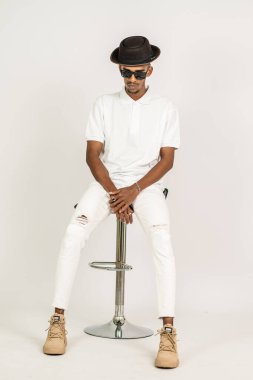 Beyaz, boş tişörtlü, şapkalı ve gözlüklü Afrikalı bir adam beyaz arka planı olan bir sandalyeye oturuyor.