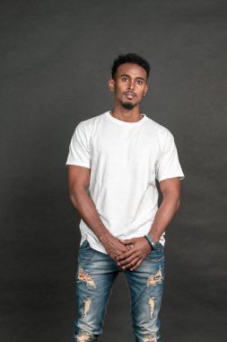 Siyah arka planda, iki elini de tutarken poz veren beyaz, boş tişört giyen genç bir Afrikalı.