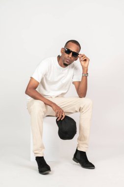 Beyaz, boş tişörtlü, şapkalı ve gözlüklü bir Afrikalı beyaz arka planda kare bir sandalyeye oturuyor.