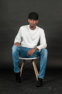 Beyaz, boş, uzun kollu gömlek ve şapka giyen yakışıklı bir adam siyah arka planda bir sandalyeye oturuyor.