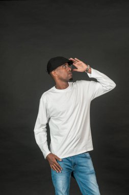 Beyaz, boş, uzun kollu bir gömlek giyen Afrikalı bir adam elini şapkasının üstüne koyup siyah arka planda poz veriyor.