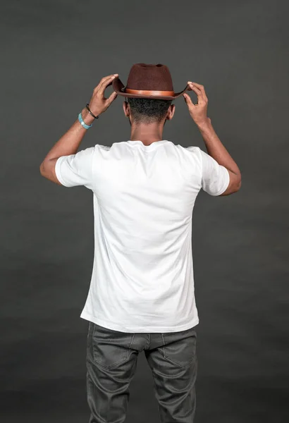 Back side image of an african man wearing white blank shirt with a hat, doing a simple pose by putting both of his hand on his hat, on the black background