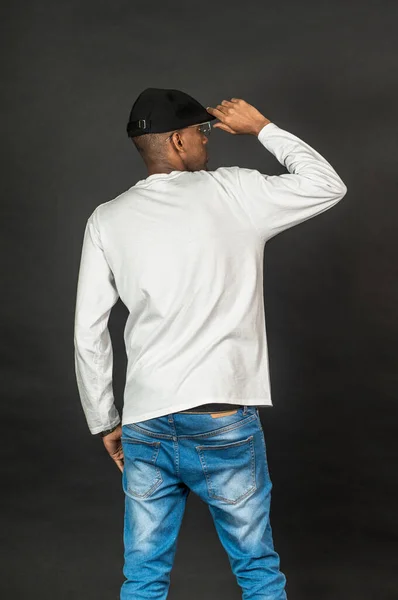 Back side image of an african man wearing white blank long sleeve shirt, doing a pose with one of his hand on his hat, on the black background