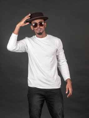 Siyah arka planda beyaz, boş, gözlüklü ve şapkalı bir Afrikalı genç bir adam elinde şapkasıyla poz veriyor.