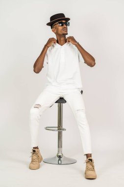 Beyaz, boş tişörtlü, şapkalı ve gözlüklü Afrikalı bir adam beyaz arka planı olan bir sandalyeye oturuyor.