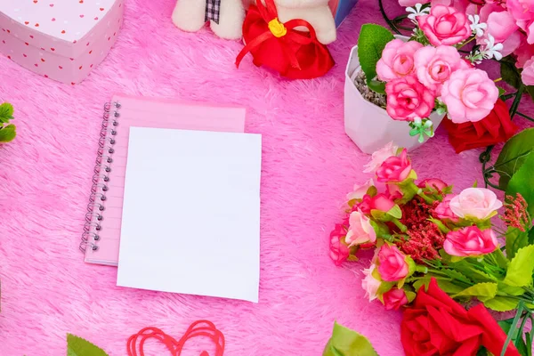 White blank notebook paper on the top of a pink covered notebook surrounded by valentine themed decorations, and a pink fluffy carpet as the background