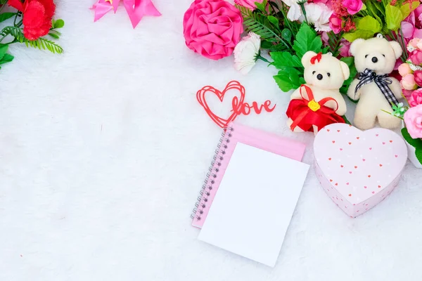 White blank notebook paper on the top of a pink covered notebook surrounded by valentine themed decorations, and a white fluffy carpet as the background